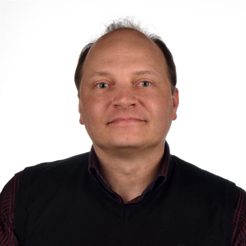 Lyhythiuksinen henkilö, jolla on tumma villapaita kauluspaidan päällä, hymyilee kameralle valkoista taustaa vasten.